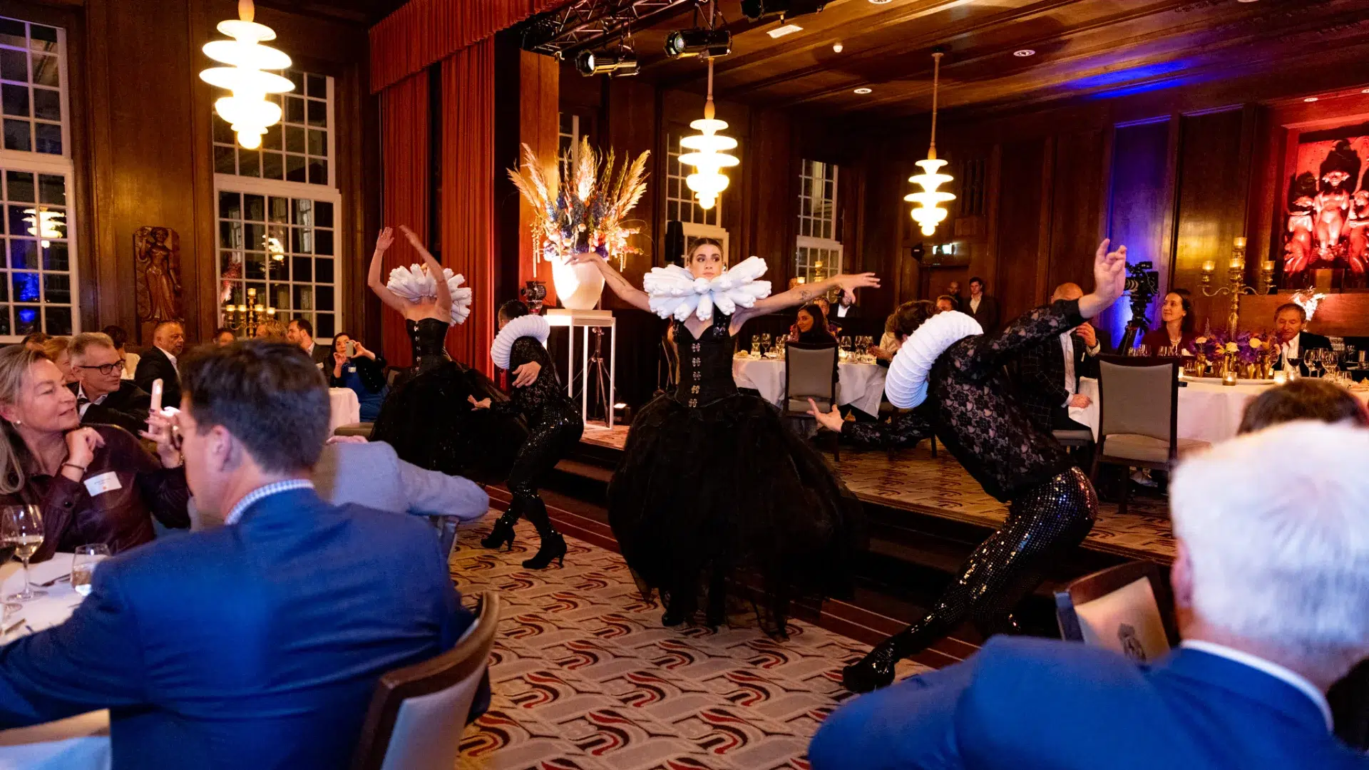 Live performers in dramatic costumes dancing among guests at a corporate gala dinner in a grand Amsterdam ballroom, the kind of entertainment that moves day two from company context into a genuine shared experience