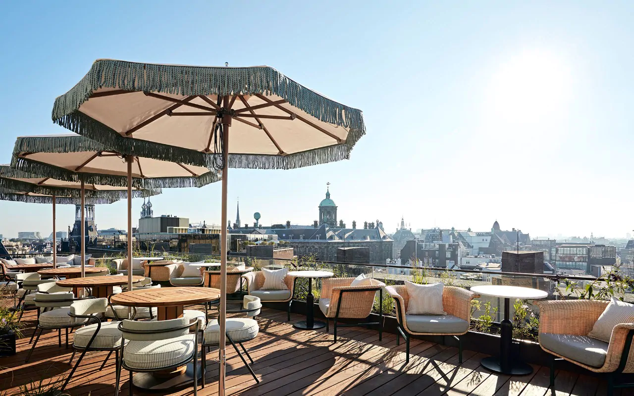 Rooftop terrace with rattan furniture and fringed parasols overlooking Amsterdam's historic skyline on a sunny day — an outdoor venue that only works when season and city calendar are confirmed together, not treated as separate planning steps