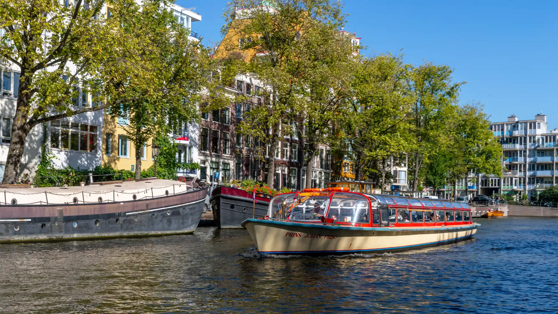 Classic canal boat on an Amsterdam waterway passing houseboats and gabled canal houses, showing how group water transfers can be built into a multi-day incentive programme as an experience in their own right