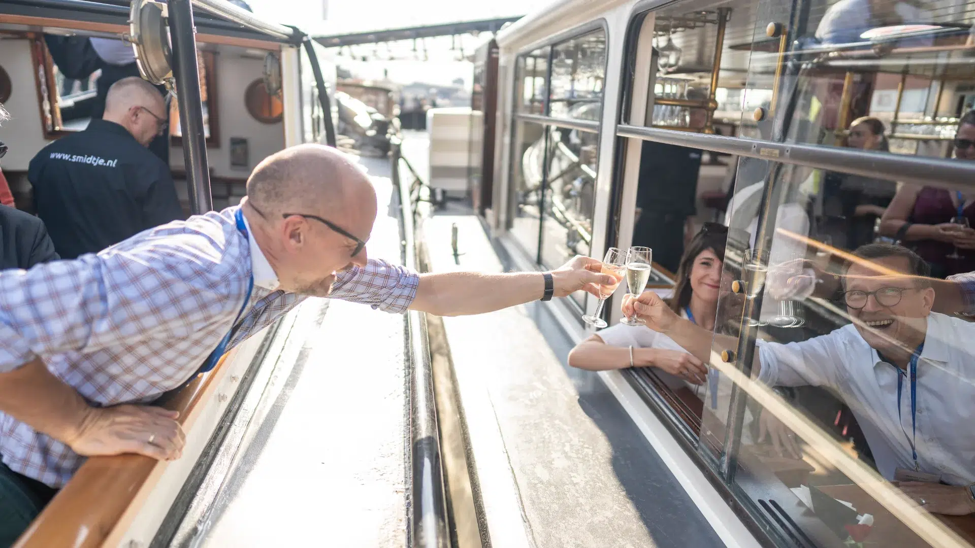 Corporate guests clinking champagne glasses through the window of a classic Amsterdam canal boat — water-based hospitality that turns a programme transfer into a social moment without adding a separate activity to the schedule