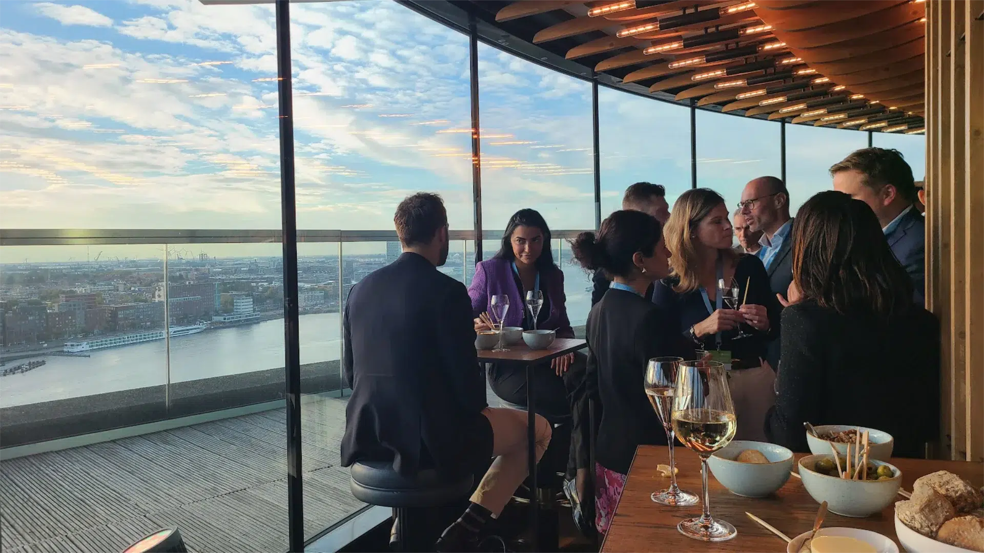 Corporate delegates in informal conversation over drinks at a panoramic Amsterdam venue overlooking the IJ harbour, showing how the right setting gives a global team room to connect beyond the structured programme