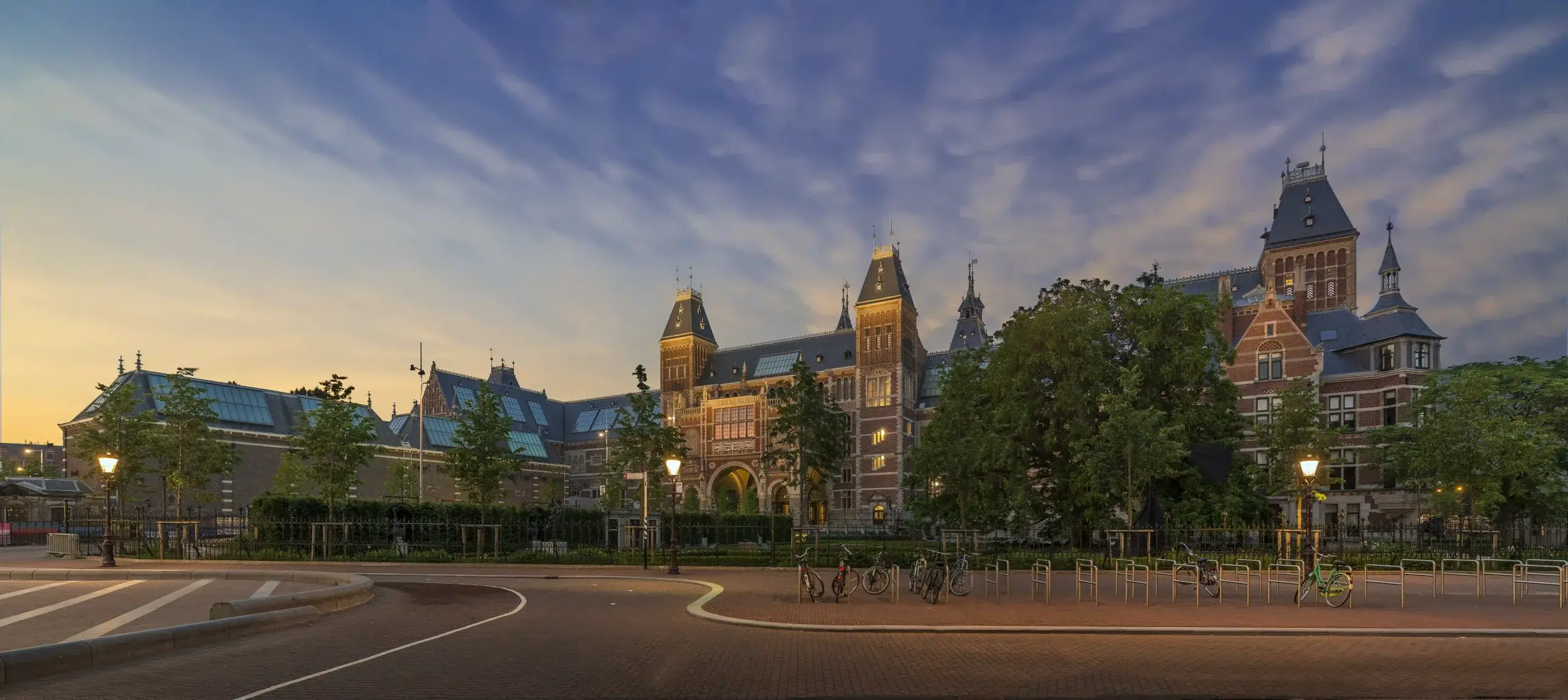 Rijksmuseum Amsterdam seen from Museumplein at dusk with street lamps lit and bicycles parked — a landmark that represents the cultural access Amsterdam offers corporate groups when the brief calls for more than a conference room